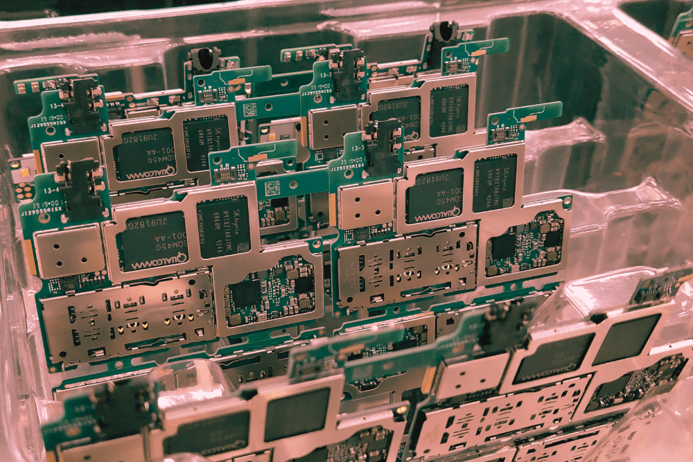 A close-up view of multiple smartphone circuit boards stacked in a plastic tray, featuring Qualcomm and SK Hynix chips.