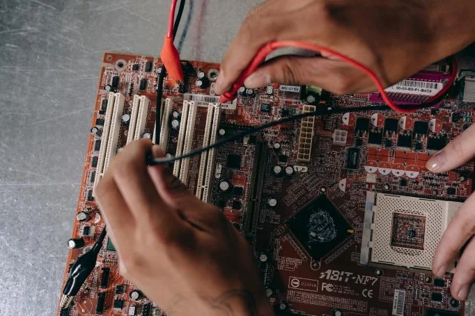 Technicians working on a red ABIT-NF7 motherboard, using testing probes to check electronic components.