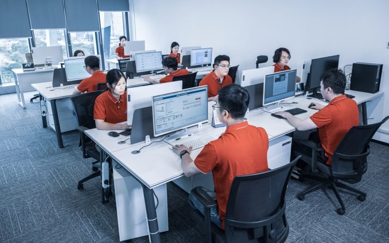 A diverse group of professionals collaborating in an office setting, each engaged with their computers and focused on tasks.